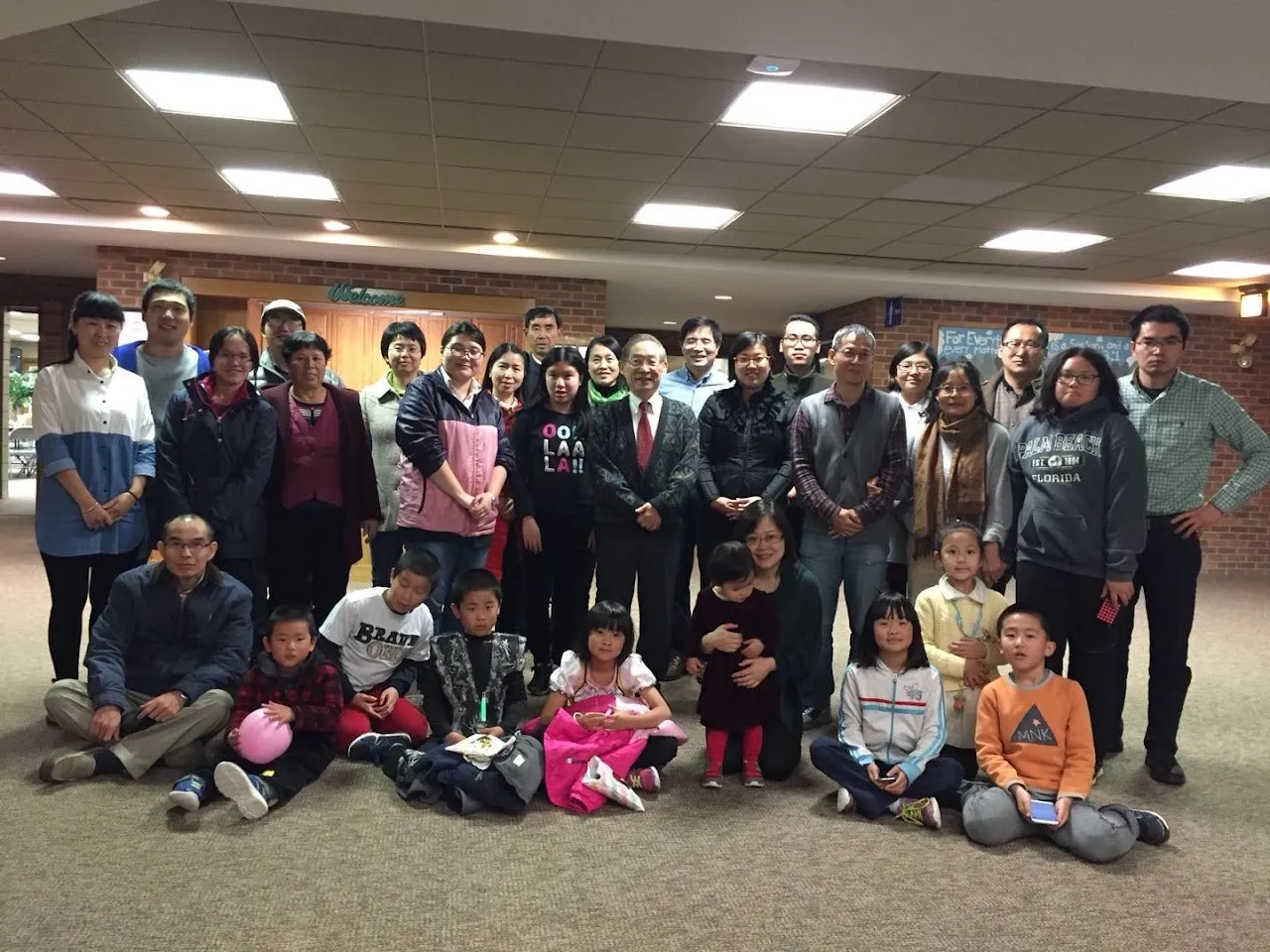 Group photo of about 35 adults and children in a church lobby during Pastor Tsu-Kung Chuang’s visit