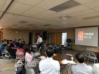A speaker presenting to an audience seated at round tables; the slide reads 平衡美满的人生